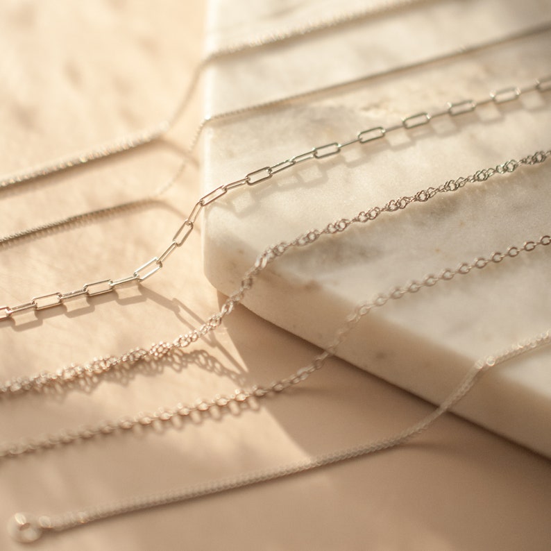 May include: A close-up of five silver chains of different styles and thicknesses, laid out on a white marble surface.