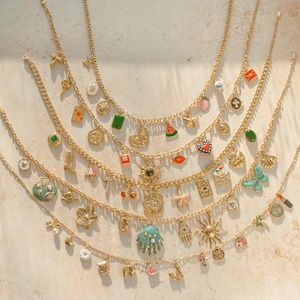 K&ouml;nnte beinhalten: Eine Sammlung von f&uuml;nf goldfarbenen Metallketten, die jeweils mit einer Vielzahl von bunten Emaille- und Metallanh&auml;ngern wie Herzen, Sternen, Muscheln, Fr&uuml;chten, Buchstaben und verschiedenen symbolischen Formen an den Gliedern versehen sind.