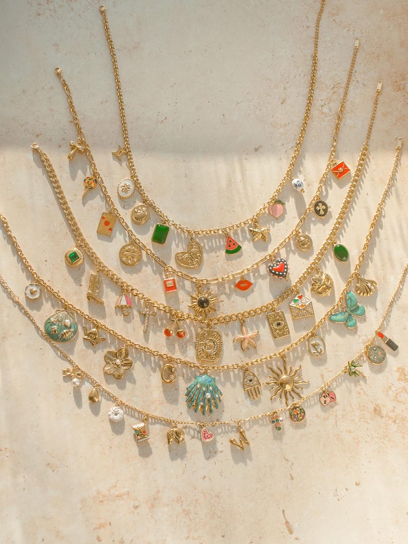 K&ouml;nnte beinhalten: Eine Sammlung von f&uuml;nf goldfarbenen Metallketten, die jeweils mit einer Vielzahl von bunten Emaille- und Metallanh&auml;ngern wie Herzen, Sternen, Muscheln, Fr&uuml;chten, Buchstaben und verschiedenen symbolischen Formen an den Gliedern versehen sind.