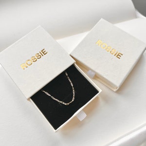 K&ouml;nnte beinhalten: Zwei wei&szlig;e Schachteln mit dem Wort "ROSSIE" in goldenen Lettern bedruckt. Eine Schachtel ist ge&ouml;ffnet und enth&auml;lt eine goldene Kettenkette auf schwarzem Hintergrund.