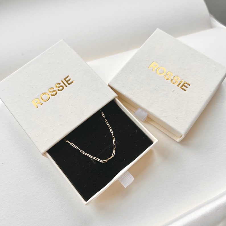 K&ouml;nnte beinhalten: Zwei wei&szlig;e Schachteln mit dem Wort "ROSSIE" in goldenen Lettern bedruckt. Eine Schachtel ist ge&ouml;ffnet und enth&auml;lt eine goldene Kettenkette auf schwarzem Hintergrund.