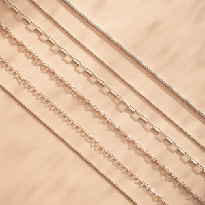May include: Five different silver chains on a beige background. The chains are arranged in a diagonal line from the top left to the bottom right of the image.