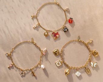 Op de afbeelding: Drie goudkleurige bedelarmbanden met diverse hangers. De bovenste armband heeft een rood hart, een klein zwart tasje en een minuscuul vliegtuigje. De linkerarmband onderaan bevat een ster, een roze hart en een parel. De rechterarmband onderaan toont een vlinder, een kruis en de letter G.