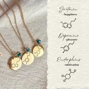 Könnte beinhalten: Drei goldene Halsketten mit runden Anhängern, die die chemischen Strukturen von Serotonin, Dopamin und Endorphinen zeigen. Jeder Anhänger hat einen kleinen grünen Edelstein. Die Halsketten liegen auf einem beigefarbenen Stoffhintergrund. Der Text auf dem Bild lautet: "Serotonin - Glück", "Dopamin - Vergnügen", und "Endorphine - Motivation".