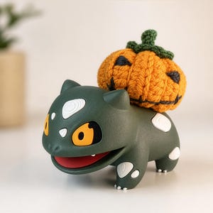 May include: A dark green creature figurine with white spots, yellow eyes, and a red mouth, featuring a crocheted orange pumpkin with a green stem and black stitched facial features resting on its back as a decorative accessory.