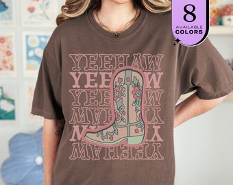 May include: A brown t-shirt with the word "YEEHAW" repeated in a pink font. A cowboy boot with floral designs is in the center. The shirt has a relaxed fit. The image also shows "8 AVAILABLE COLORS" in a purple box.