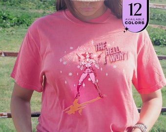 May include: A coral-colored t-shirt featuring a graphic of a cowgirl holding two guns, with the text "THE HELL I WON'T". The design includes star accents and a shadow of the cowgirl. A small graphic indicates 12 available colors.