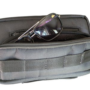 May include: Black sunglasses with a silver frame are placed inside a black pouch with a zip closure. The pouch has a textured surface and a small front pocket. The pouch is designed to hold small items, such as glasses.