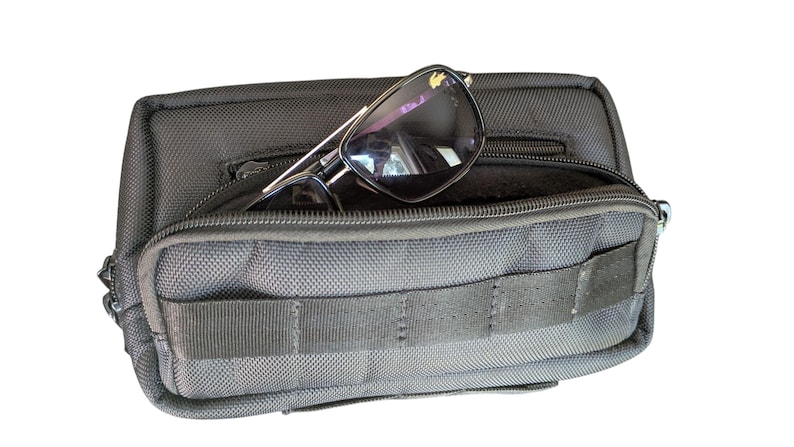 May include: Black sunglasses with a silver frame are placed inside a black pouch with a zip closure. The pouch has a textured surface and a small front pocket. The pouch is designed to hold small items, such as glasses.