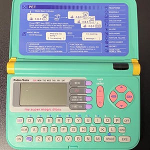 May include: A mint green Radio Shack My Super Magic Diary electronic organiser with a QWERTY keyboard, a digital screen, and pink navigation buttons. The device features a flip-top lid with printed instructions and a list of functions.