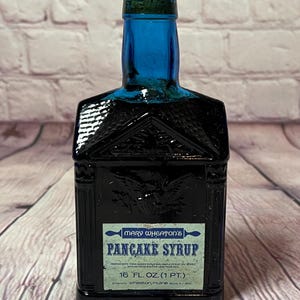 May include: A vintage, dark blue glass bottle of Mary Wheaton's Pancake Syrup. The bottle has a unique architectural shape and a cork stopper. The label reads "Pancake Syrup" and "16 fl. oz. (0.47 l)".