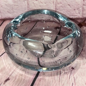 Peut inclure: Un bol rond en verre transparent avec une légère teinte bleu clair. Le bol a une large ouverture et une série de petites bulles incrustées dans le verre. Le bord du bol est incurvé, créant une forme unique.