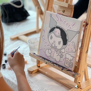 May include: A person paints a canvas with a paintbrush. The canvas features a cartoon character with a skull and crossbones, a pink bow, and the words "Sweet!" and "Oot!" The canvas is on an easel.