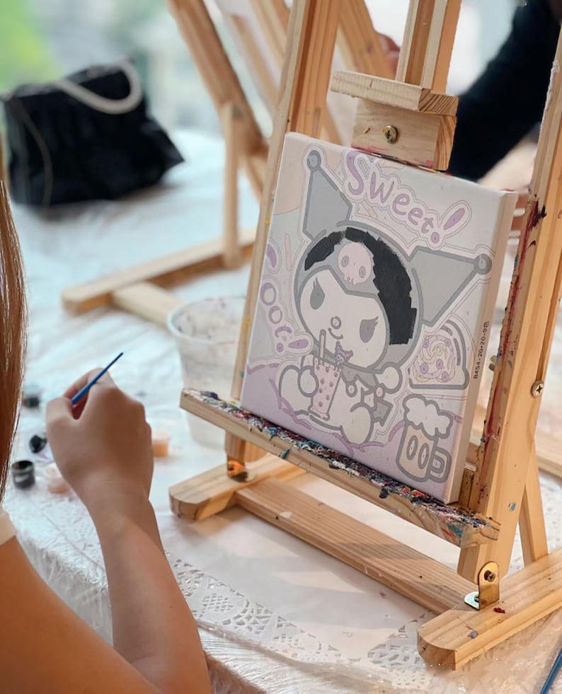 May include: A person paints a canvas with a paintbrush. The canvas features a cartoon character with a skull and crossbones, a pink bow, and the words "Sweet!" and "Oot!" The canvas is on an easel.