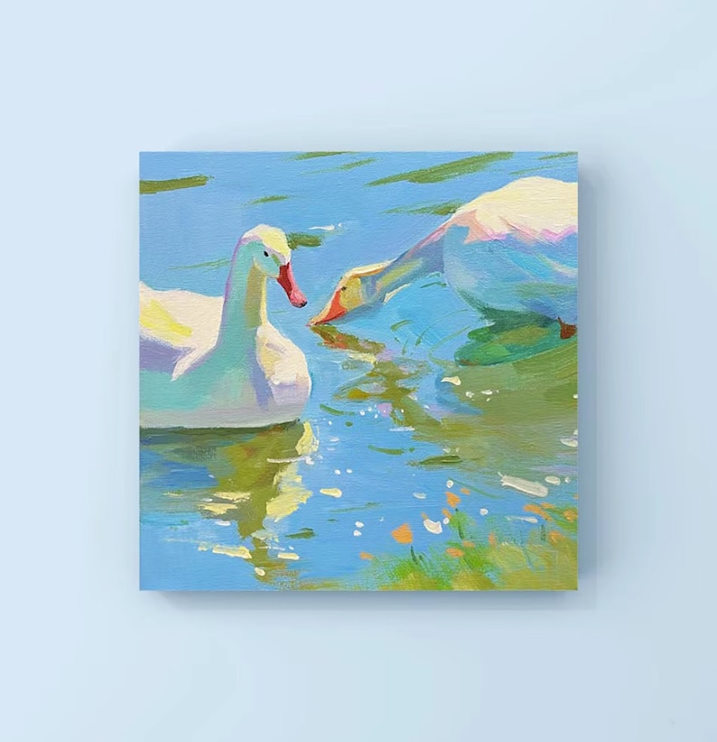 May include: Two white geese swimming in a blue pond with green grass and yellow flowers. The geese are facing each other, with one looking directly at the viewer.