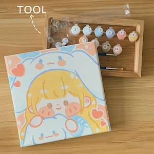 May include: A paint by numbers kit featuring a cartoon illustration of a girl with blonde hair wearing a white bunny hat and holding a white bunny. The kit includes a canvas, paint pots, and brushes.