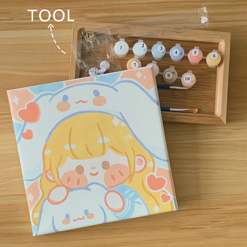 May include: A paint by numbers kit featuring a cartoon illustration of a girl with blonde hair wearing a white bunny hat and holding a white bunny. The kit includes a canvas, paint pots, and brushes.