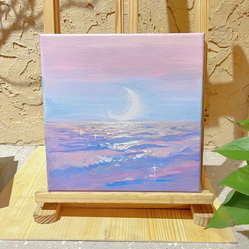 May include: A painting of a crescent moon over a purple and blue landscape with white stars. The painting is on a small wooden easel.