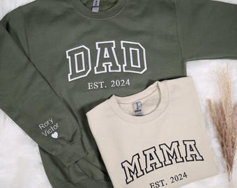 May include: Two sweatshirts, one green and one beige, with the words "DAD EST. 2024" and "MAMA EST. 2024" printed on them. The green sweatshirt has the words "Rory Victor" and a heart on the sleeve.