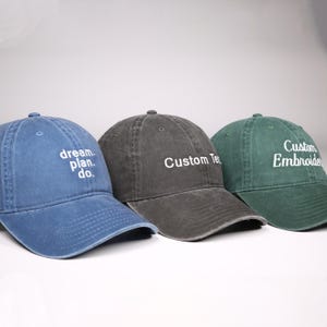 May include: Three baseball caps in blue, charcoal, and green. The blue cap has the text "dream. plan. do." in white. The charcoal cap says "Custom Text" and the green cap says "Custom Embroidered" in white. All caps have a curved brim.