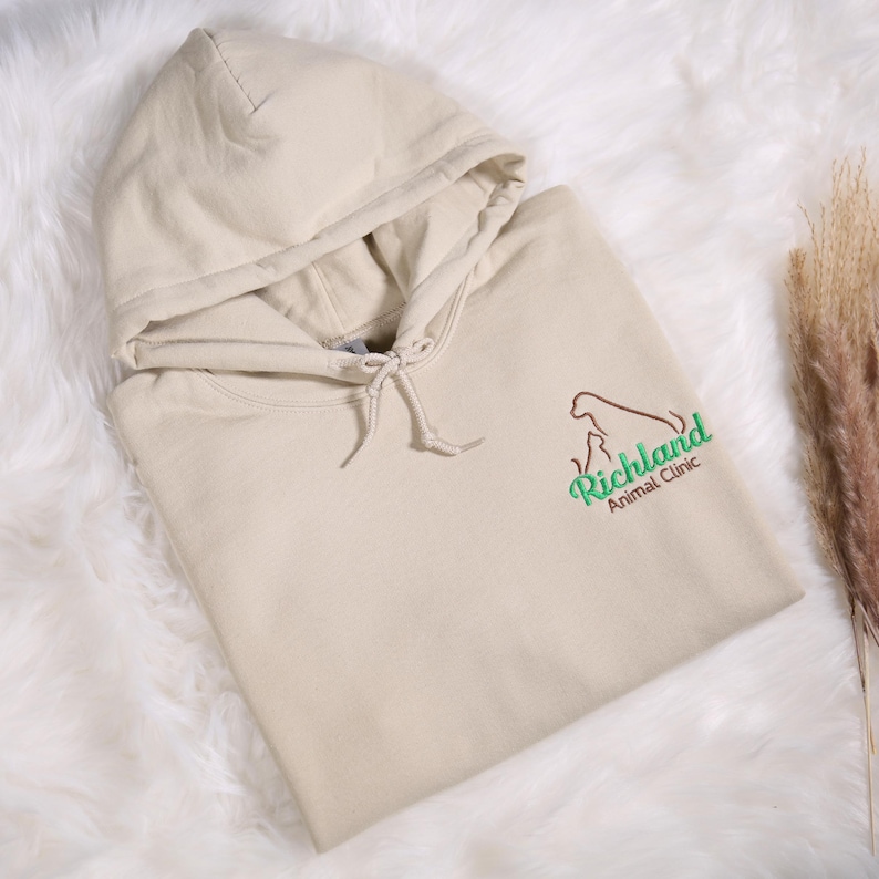 May include: A beige hooded sweatshirt with a drawstring and a front pocket. The sweatshirt features the logo for Richland Animal Clinic in brown and green. The garment is folded on a white, fluffy surface.