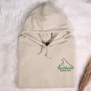 May include: A beige hoodie with a drawstring hood. The front of the hoodie features a logo of a dog and cat with the words "Richland Animal Clinic" in green and brown.