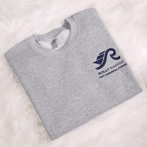 May include: A heather gray sweatshirt with a navy blue logo that reads "Rolly Marine Fort Lauderdale, Florida." The sweatshirt is folded and has a crew neck.