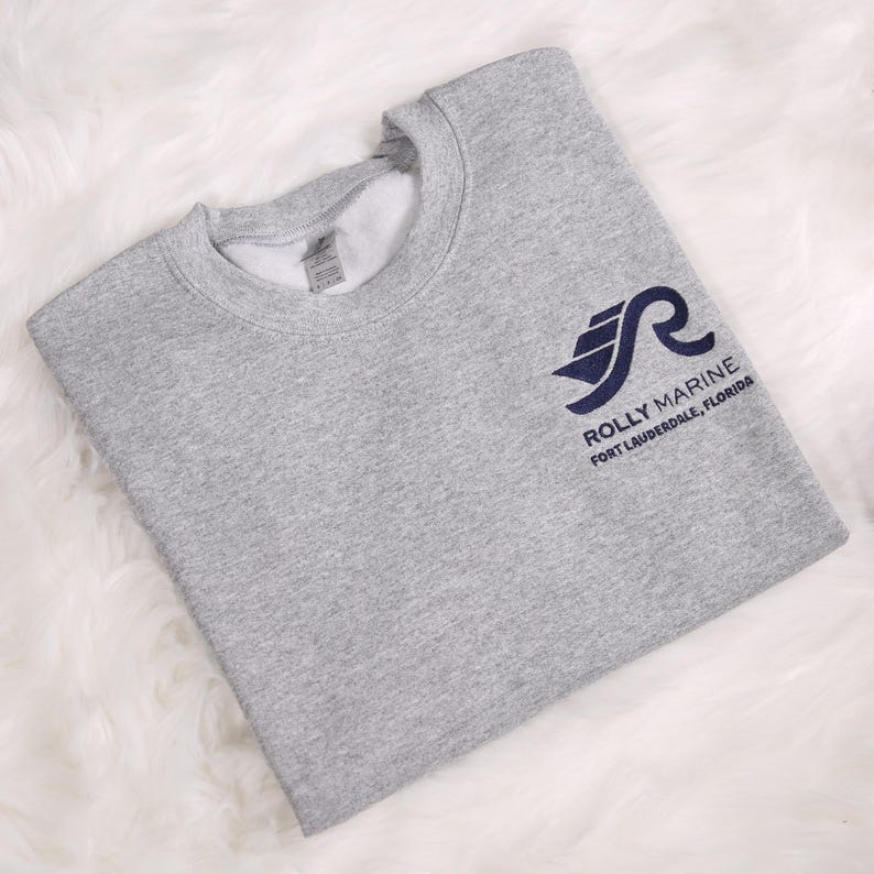 May include: A heather gray sweatshirt with a navy blue logo that reads "Rolly Marine Fort Lauderdale, Florida." The sweatshirt is folded and has a crew neck.