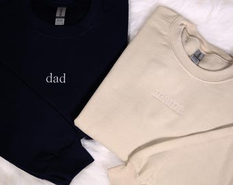 Puede incluir: Dos sudaderas, una azul marino y otra beige, con las palabras "papá" y "mamá" bordadas.