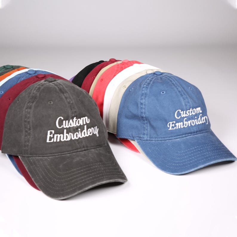 May include: A collection of baseball caps in various colors, including gray, blue, red, and black. Each cap features the embroidered text "Custom Embroidery" in white. The caps are arranged in a row, showcasing their different hues.