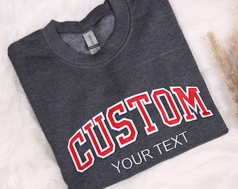 May include: A dark grey sweatshirt with a red embroidered arch that says "CUSTOM" and white text below that says "YOUR TEXT".