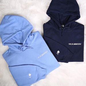 Peut inclure: Deux sweat-shirts à capuche, l'un bleu clair et l'autre bleu marine, avec des chiffres romains brodés en blanc et un symbole de cœur. Le sweat-shirt bleu clair porte la date IV.XI.MMMXIV et le sweat-shirt bleu marine porte la date VII.XI.MMMXIV.