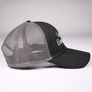 May include: Black trucker hat with a gray mesh back and a curved brim. The hat features white embroidered text on the front panel. A small black button is on top of the hat.