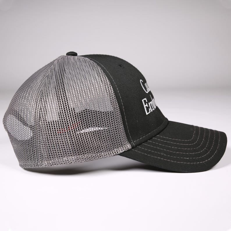 May include: Black trucker hat with a gray mesh back and a curved brim. The hat features white embroidered text on the front panel. A small black button is on top of the hat.