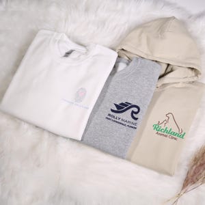 May include: Four sweatshirts and hoodies in white, gray, and tan. The white sweatshirt has a fingerprint design and the text "Therapy Solutions". The gray sweatshirt has a boat and wave logo with the text "Rolly Marine Fort Lauderdale, Florida". The tan hoodie has a dog logo with the text "Richland Animal Clinic".