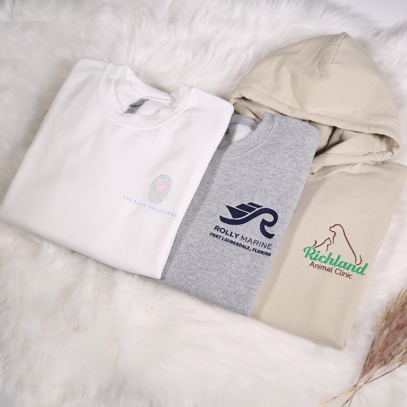 May include: Four sweatshirts and hoodies in white, gray, and tan. The white sweatshirt has a fingerprint design and the text "Therapy Solutions". The gray sweatshirt has a boat and wave logo with the text "Rolly Marine Fort Lauderdale, Florida". The tan hoodie has a dog logo with the text "Richland Animal Clinic".
