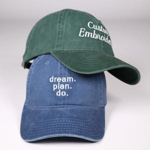 May include: Two baseball caps, one green and one blue, stacked on top of each other. The green cap has "Custom Embroider" embroidered in white. The blue cap has "dream. plan. do." embroidered in white.