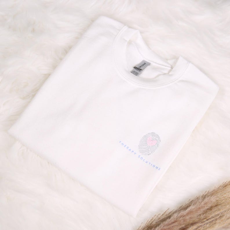 May include: A white sweatshirt with a crew neck. The sweatshirt features a small graphic of a fingerprint with a heart in the center and the words "THERAPY SOLUTIONS" in a light blue font. The sweatshirt is folded neatly on a white, fluffy surface.