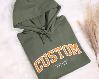 May include: A green hooded sweatshirt with the word "CUSTOM" in orange letters with a white outline. The text "TEXT" is below the word "CUSTOM".