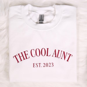 May include: White sweatshirt with red embroidered text that reads "The Cool Aunt Est. 2023".