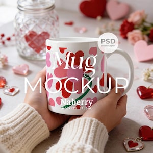 May include: White ceramic mug with a floral heart design in shades of red and pink. The mug has the words "Mug MOCKUP" printed on it, along with the name "Naberry" and "MOCKUPS STUDIO". The mug is held by a person wearing a cream-colored sweater.