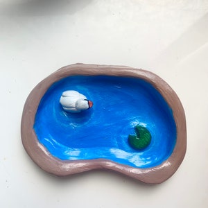 Puede incluir: Un pequeño estanque de cerámica con un cisne blanco y un nenúfar verde. El estanque está pintado de azul y los bordes son marrones.