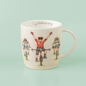 May include: Cream ceramic mug with a handle, featuring a cyclist design. The mug has the word "Cycletherapy" written on it. The illustration shows three cyclists, with one cyclist raising their arms in victory. The cyclists are wearing helmets and riding bicycles.