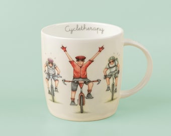 May include: Cream-colored ceramic mug with a handle, featuring a cyclist design. The mug has the word "Cycletherapy" written on it. The illustration shows three cyclists, with one cyclist raising their arms in victory. The cyclists are wearing helmets and riding bicycles.