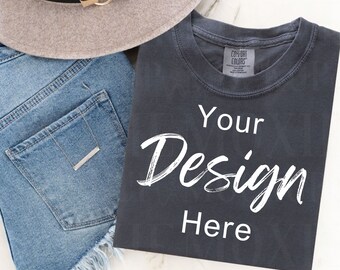 May include: A dark gray t-shirt with the text "Your Design Here" in white. The t-shirt is folded and laid on a white surface next to a pair of blue denim shorts and a brown fedora.