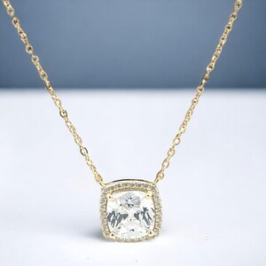 May include: A gold chain necklace with a large, square-shaped pendant featuring a clear, faceted gemstone surrounded by smaller, clear gemstones.