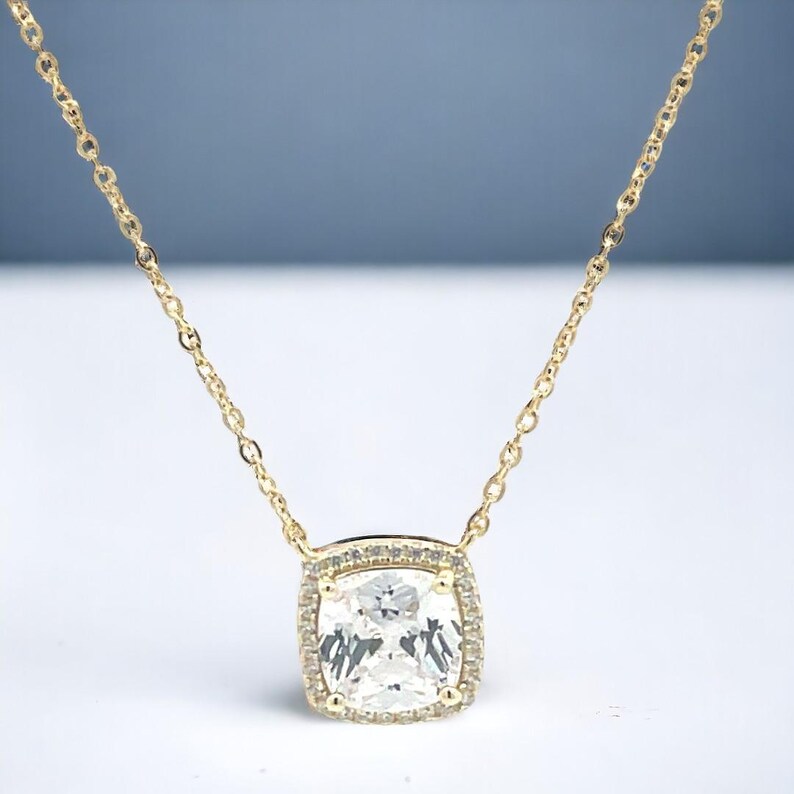 May include: A gold chain necklace with a large, square-shaped pendant featuring a clear, faceted gemstone surrounded by smaller, clear gemstones.