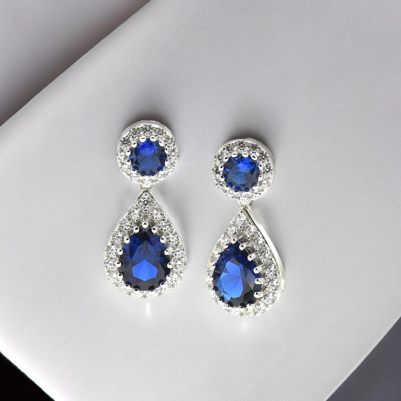 Pode incluir: Um par de brincos de prata com pedras de safira azul em forma de l&aacute;grima, cercadas por pedras menores e transparentes.
