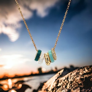May include: Gold-toned necklace featuring five decorative rings. The rings are in shades of teal, white, and gold, with some adorned with crystals and star patterns. The necklace is displayed against a blurred backdrop of a sunset and sky.