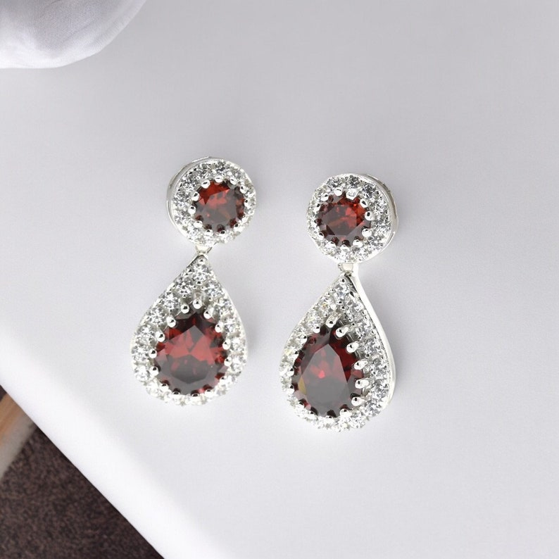 Pode incluir: Um par de brincos de prata com pedras vermelhas e transparentes. Os brincos t&ecirc;m um design em forma de l&aacute;grima com uma pedra redonda menor na parte superior.